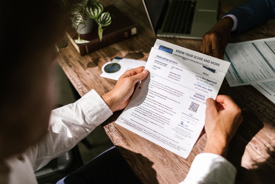 Person holding and reading a printed credit score report at a wooden desk, with a laptop, papers, a cup, and a small plant nearby.