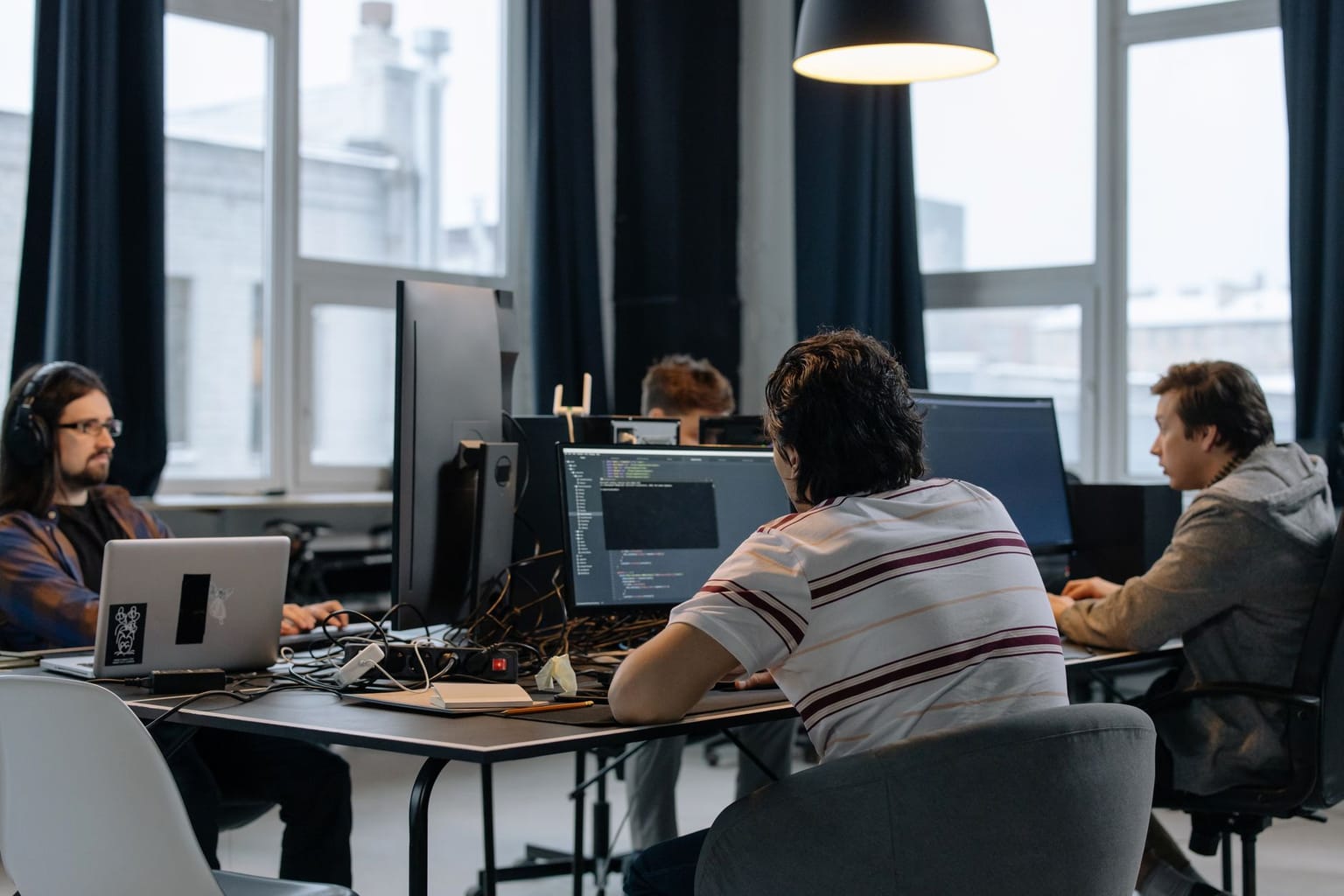 Three people sit at desks with multiple monitors, coding in an office with large windows and dark curtains. Cables and laptops are visible on the
