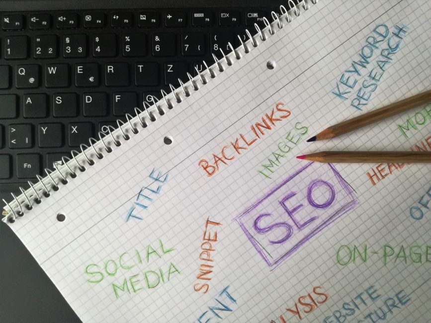 A spiral notebook with SEO-related words written in colored pencil rests on a laptop keyboard beside two colored pencils