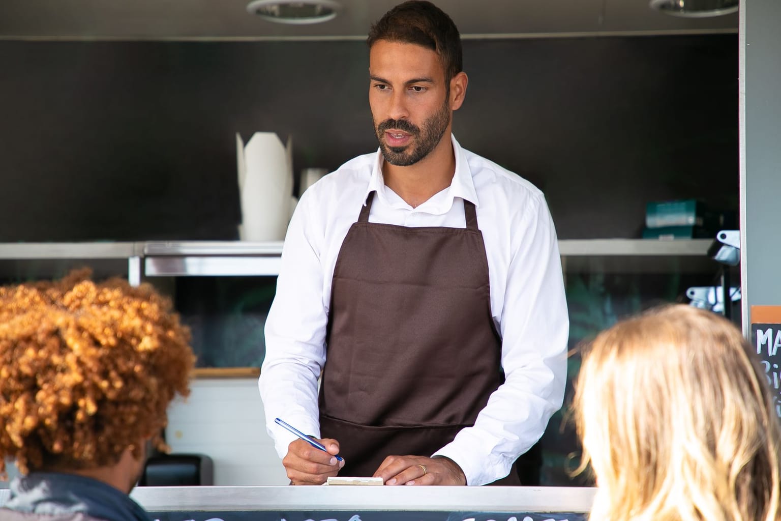 A man wearing a white shirt and brown apron stands at a counter, holding a pen and notepad, taking orders from customers in front of a food truck