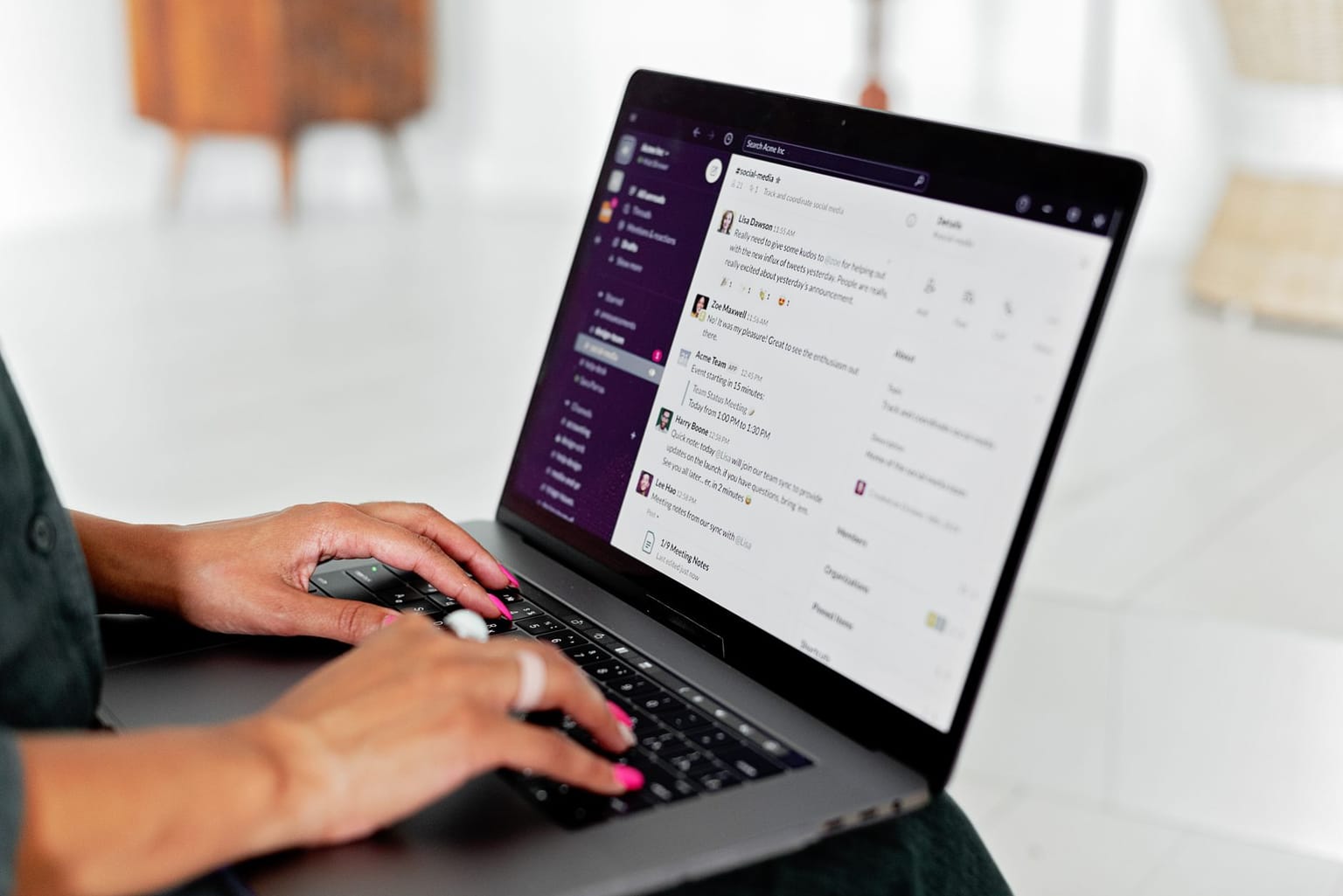A person with pink nail polish types on a laptop, viewing a Slack workspace with messages and channels displayed on the screen