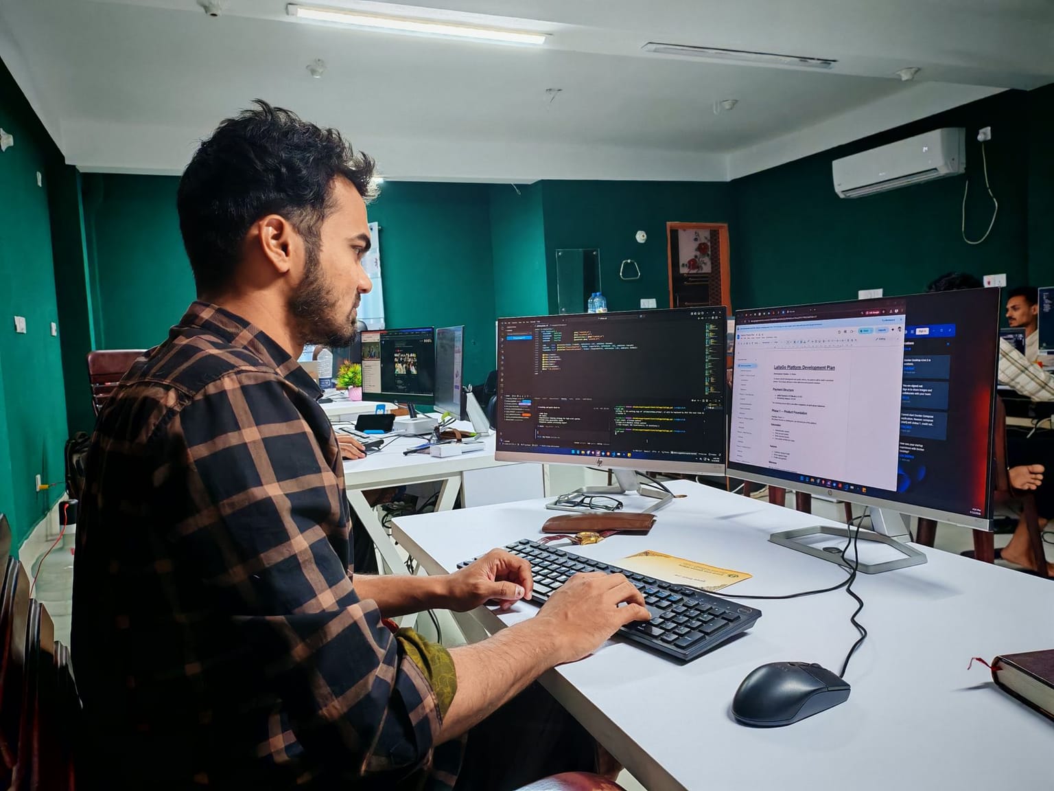 A man sits at a desk in an office, working on a computer with two monitors displaying code and a webpage