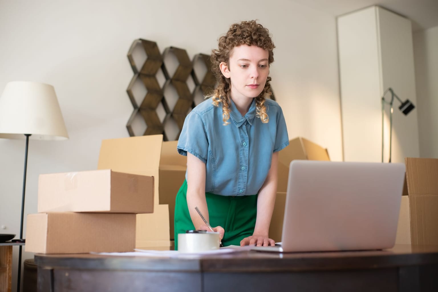A person stands at a table with a laptop, surrounded by cardboard boxes, writing notes. The room has a lamp and shelves in the background