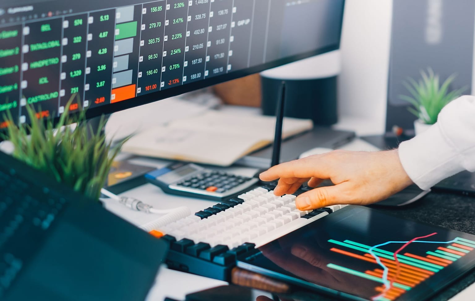 A person operates a keyboard at a desk with two monitors displaying stock market data and colorful graphs