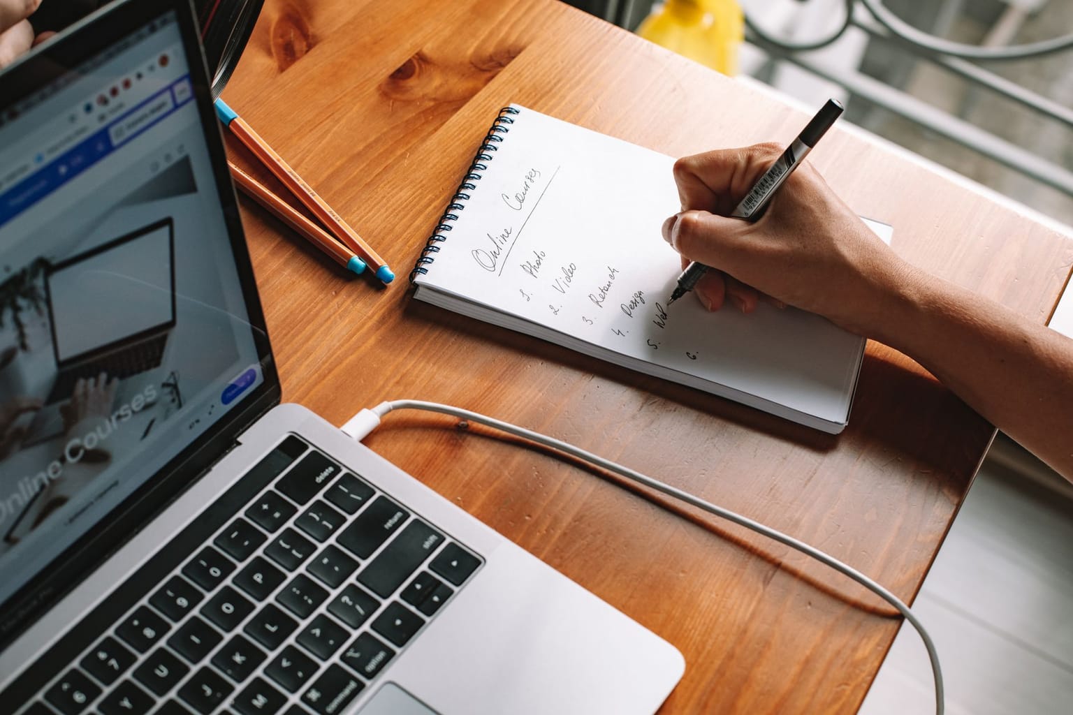 Person writing a to-do list in a notebook next to a laptop and some pens on a wooden table. The laptop screen shows an online course website
