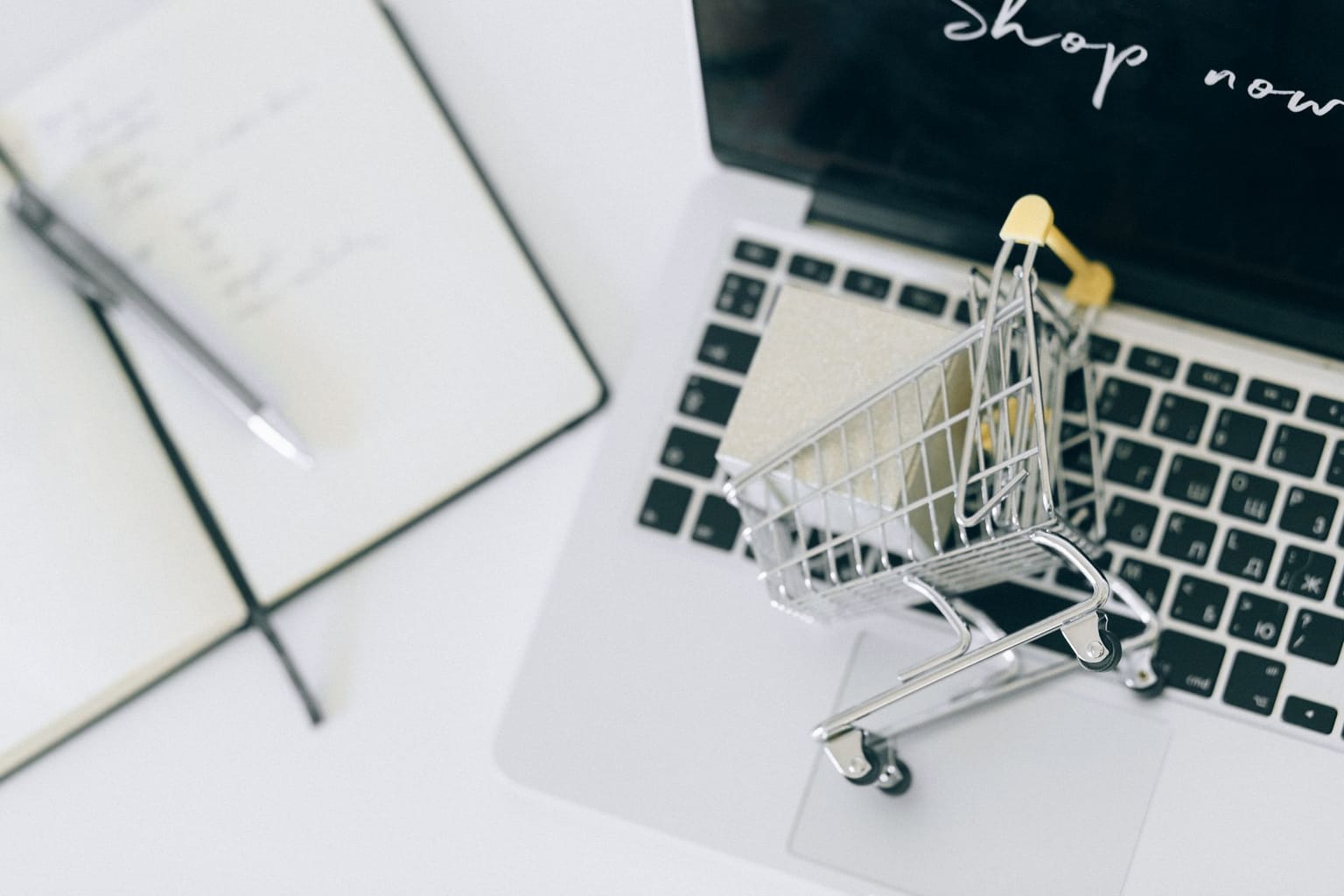 A small shopping cart sits on a laptop keyboard next to an open notebook and pen, symbolizing buying software online or finding a lifetime deal