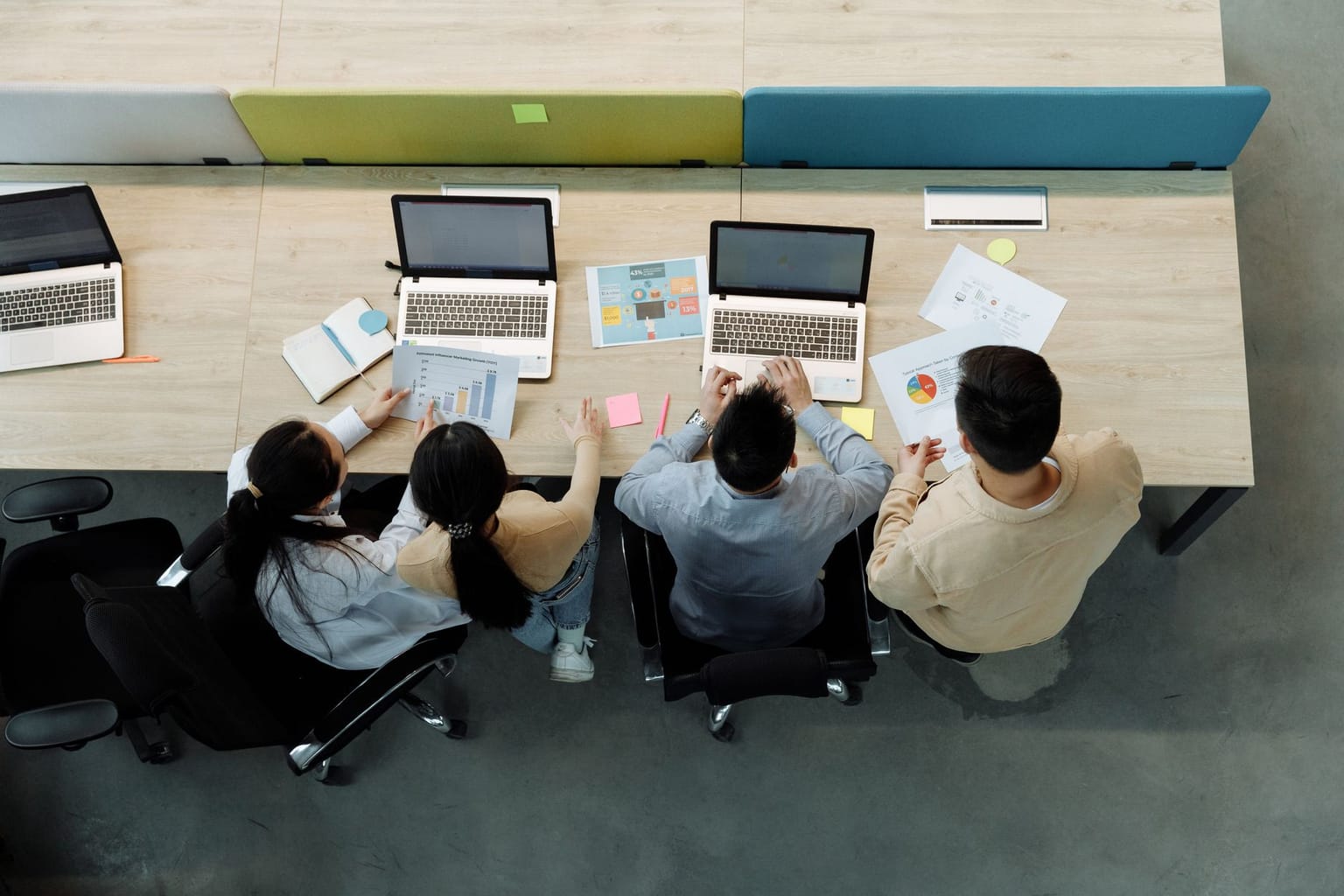Four startup team members sit at a shared desk, working on laptops with documents, charts, sticky notes